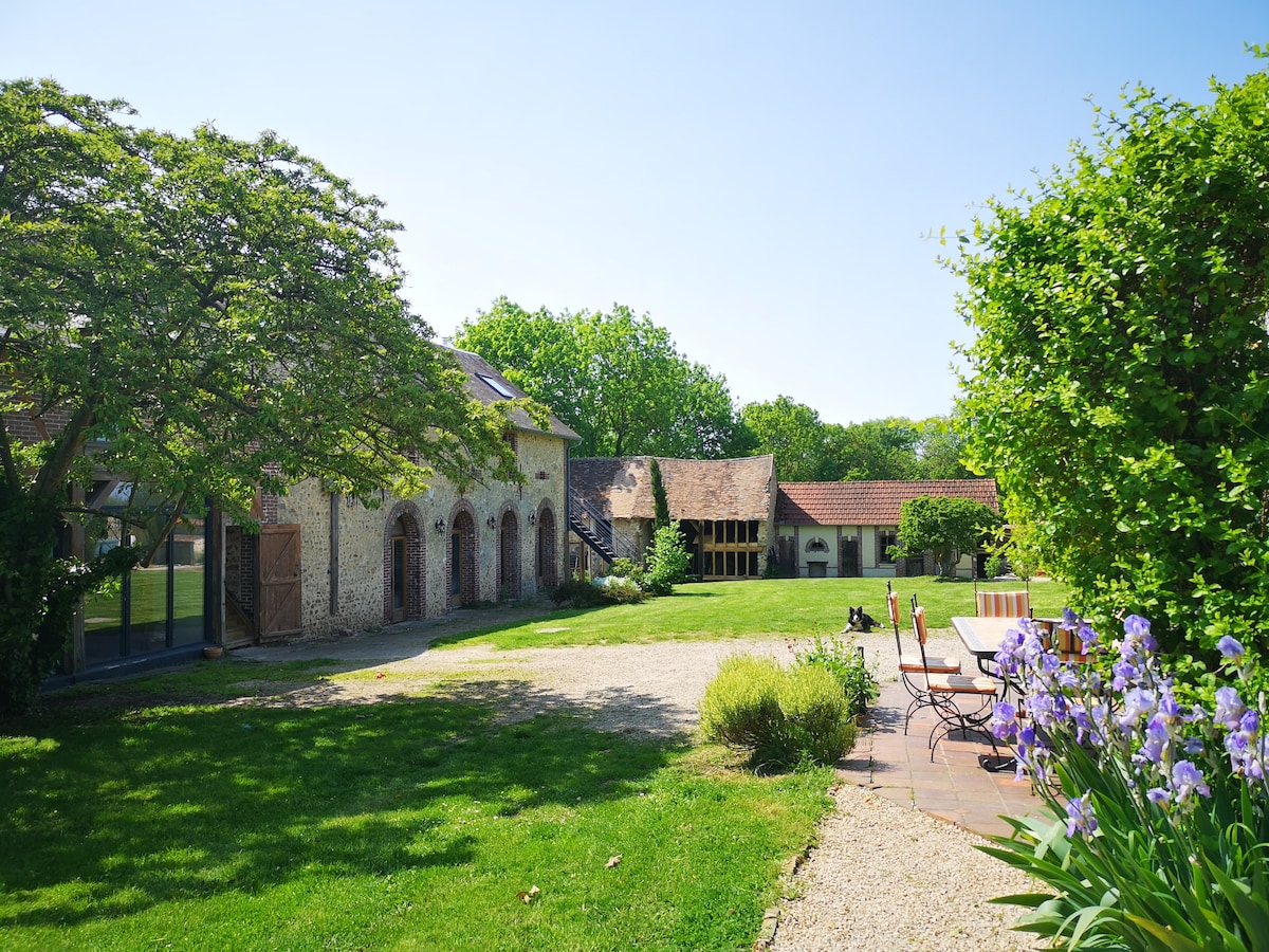 The outdoor area is surrounded by lush greenery, featuring a well-maintained lawn and flowering plants. Several stone buildings, characteristic of the property's history, are visible, along with a seating area containing a small table and chairs, providing a peaceful place for relaxation.
