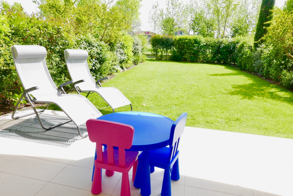 A vibrant outdoor space is presented with a lush green lawn framed by hedges. Two lounge chairs are positioned nearby, offering a place to relax, while a small blue and pink table with matching chairs is situated on the patio, ideal for children's activities.