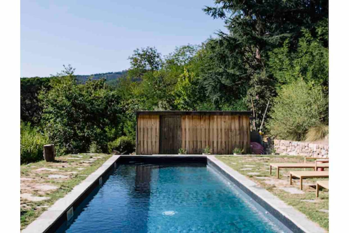 An outdoor swimming pool is surrounded by lush greenery, with a wooden shed in the background. Lounge chairs are positioned nearby, providing a space for relaxation under clear blue skies. The tranquil setting emphasizes the peacefulness of the natural environment.