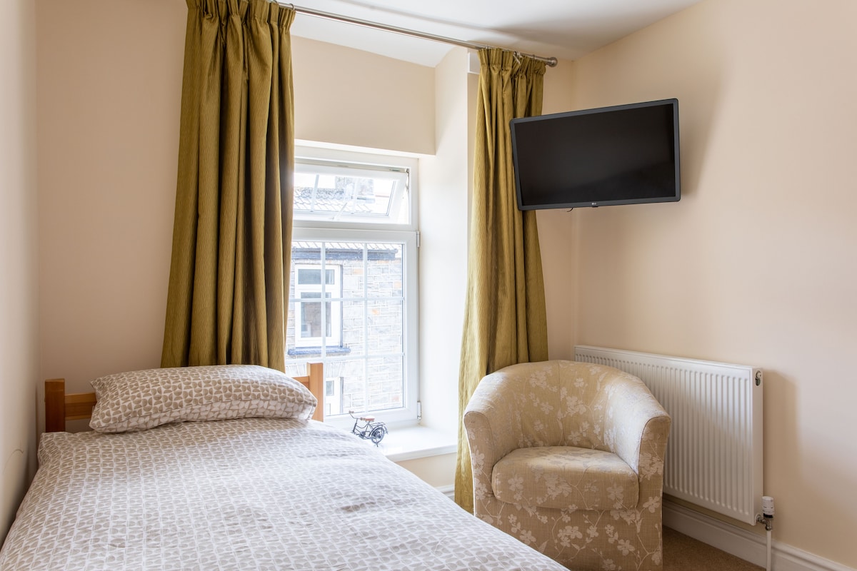 A simple bedroom is presented with a spacious bed covered in a patterned duvet. A small armchair is positioned near the window, which is framed by light-colored curtains. A wall-mounted television is visible, and the room is warmed by a radiator beneath the window.
