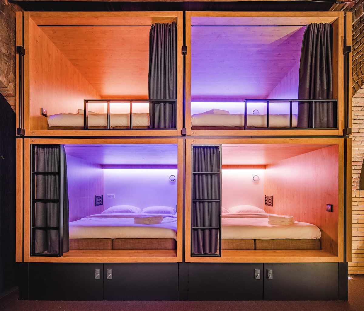 Four sleeping pods are arranged in a grid, designed for privacy and comfort. Each pod features a spacious bed with cozy bedding, accentuated by soft LED lighting. Dark curtains can be drawn for added seclusion, while outlets are visible on the lower panel.