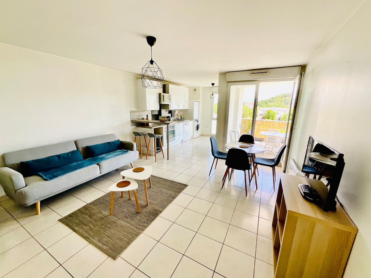 The bright living area features a gray sofa and two small accent tables atop a woven rug. A dining table with four chairs is positioned near large glass doors, which lead to a balcony with scenic views. The open kitchen is equipped with modern appliances.
