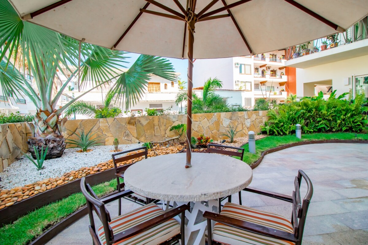 An outdoor seating area features a round table with four wooden chairs under a large umbrella. Surrounding the area are tropical plants and decorative stones, creating a lush and inviting environment.