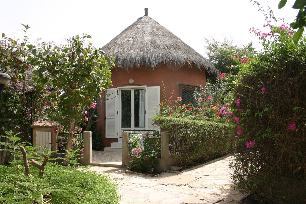 A charming villa with a thatched roof is surrounded by lush greenery and vibrant flower bushes. The entrance features a paved pathway leading to white shutter doors, inviting light into the interior. The outdoor space offers a serene garden atmosphere.
