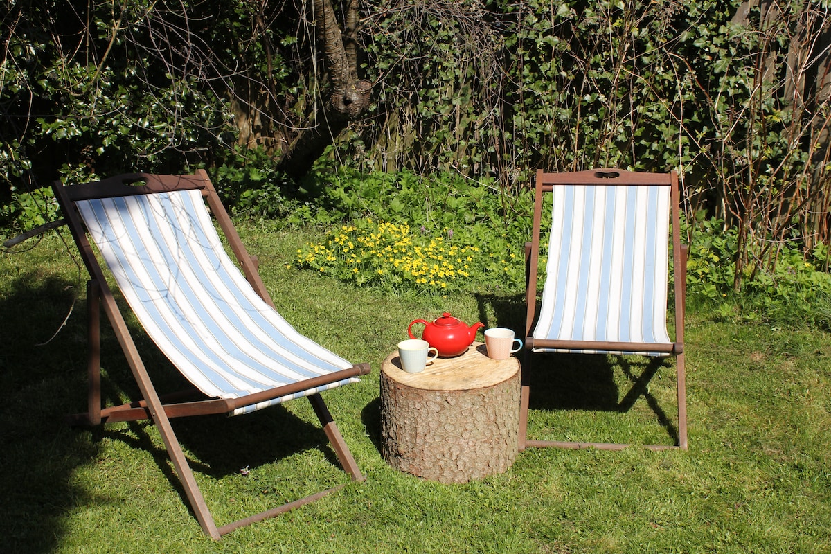 Two wooden lounge chairs with striped fabric are positioned around a round tree stump serving as a table. A red kettle and two white cups are placed on the stump, surrounded by a lush green yard filled with blooming flowers and greenery.