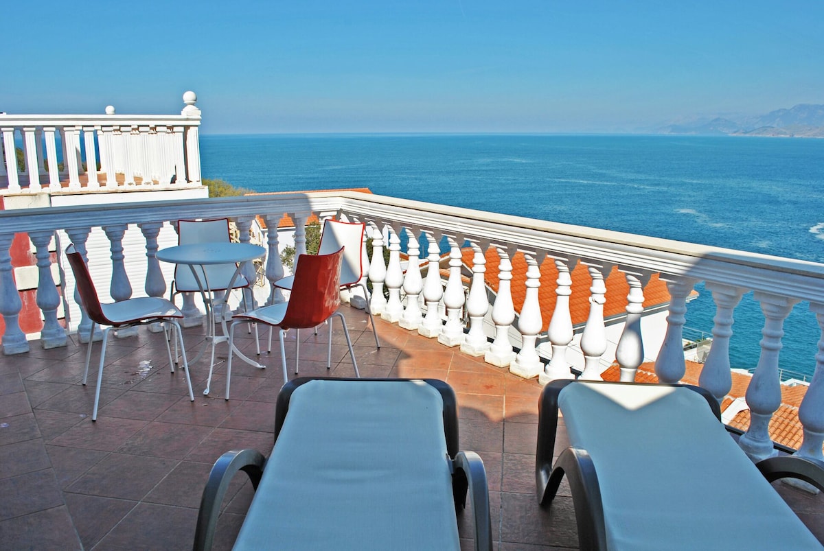A terrace area features two lounge chairs and a small dining table with four chairs, all set against a backdrop of expansive sea views. The white railing outlines the space, contributing to a serene atmosphere with clear blue water in the distance.