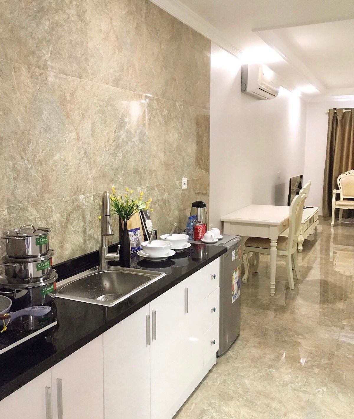 A modern kitchen area features sleek white cabinetry with a black countertop. Stainless steel cooking utensils are neatly arranged beside a small sink. A compact refrigerator is placed near a dining area set with white chairs, while subtle decorations add a touch of elegance.