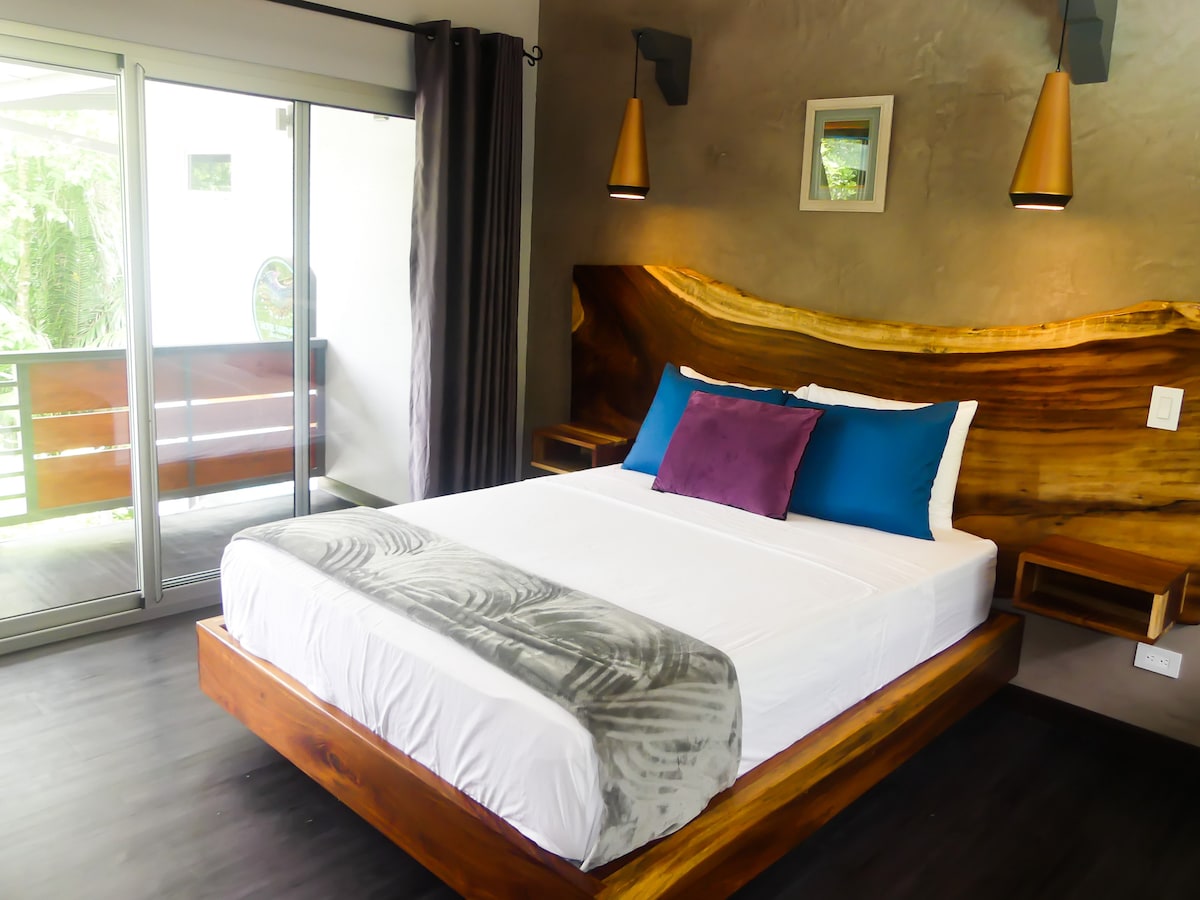 A cozy bedroom features a spacious bed with a white duvet and decorative pillows in shades of blue and purple. Smooth wooden accents frame the bed, while large sliding glass doors allow natural light and provide access to a private balcony.