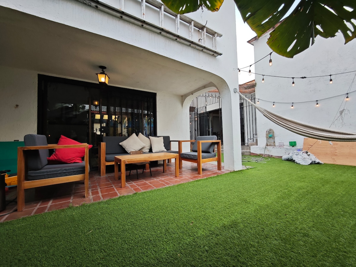 A shaded outdoor seating area is featured, with comfortable wooden chairs and cushions arranged around a low table. A hammock is visible hanging nearby, and fresh green grass carpets the ground. String lights create a bright overhead ambiance, enhancing the inviting atmosphere.