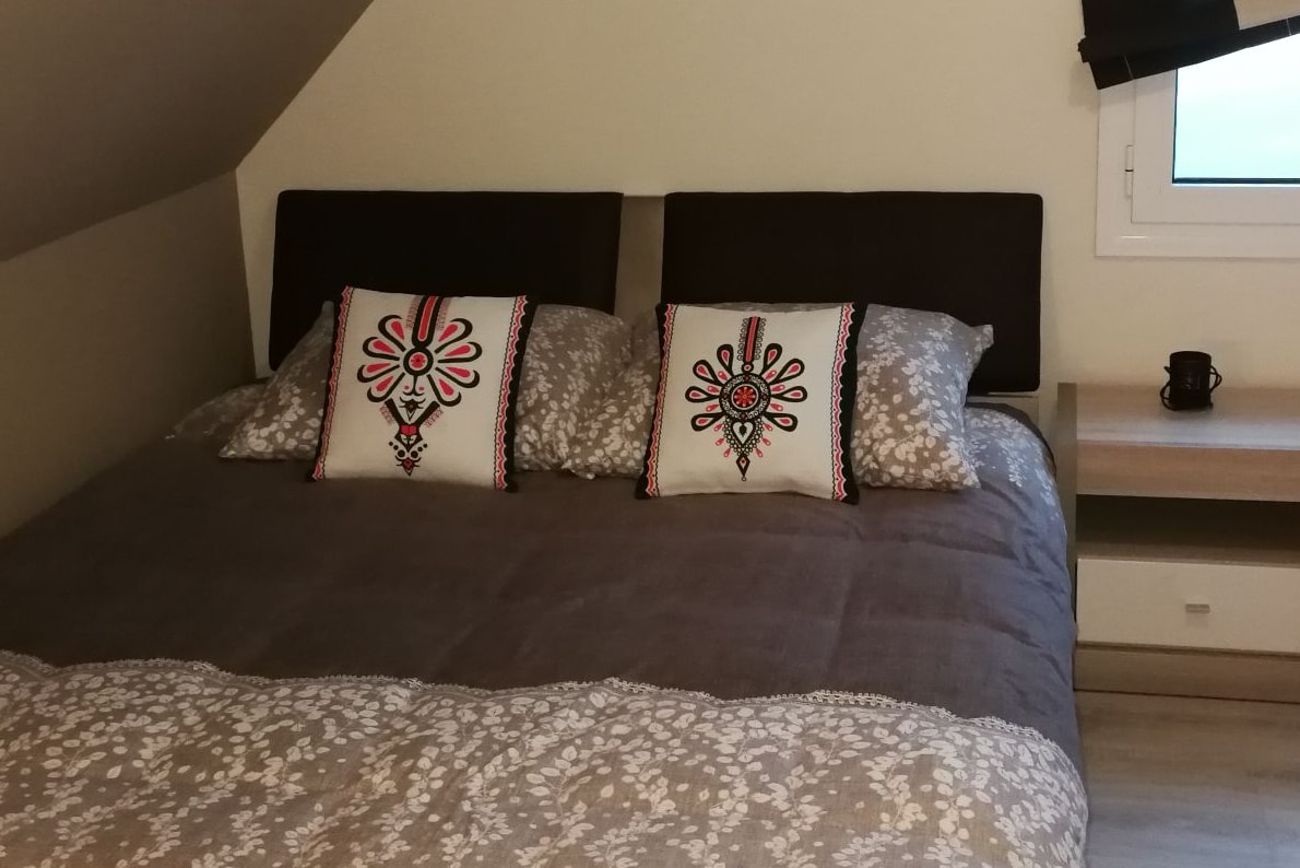 A spacious bed is adorned with decorative pillows featuring floral patterns. The neutral bedding complements the wood accents of the room. A bedside table with a cup adds a functional touch, while natural light filters through a nearby window.