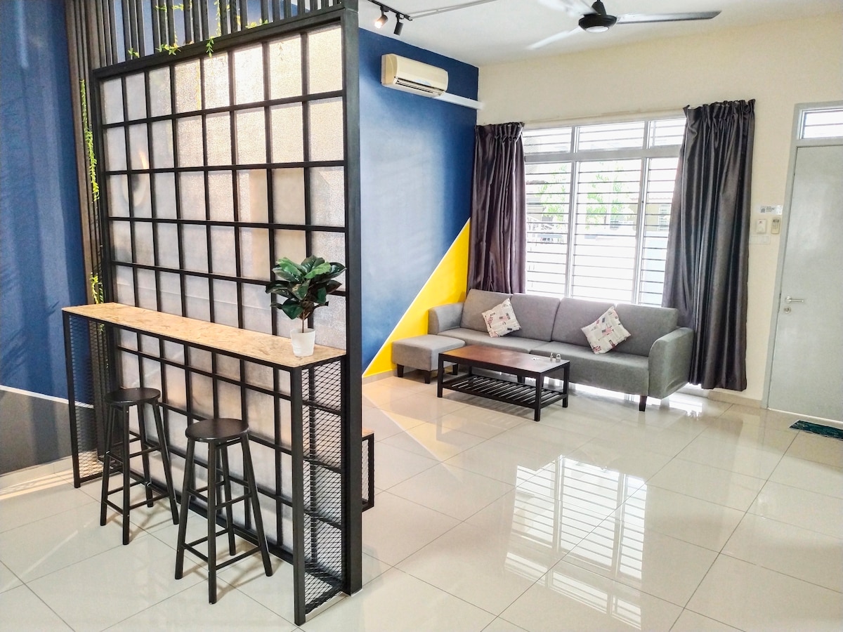 An open living area is divided by a modern screen, featuring a bar-like table with two stools. A grey sofa and a black coffee table are arranged nearby, with natural light filtering in through large windows adorned with light curtains.