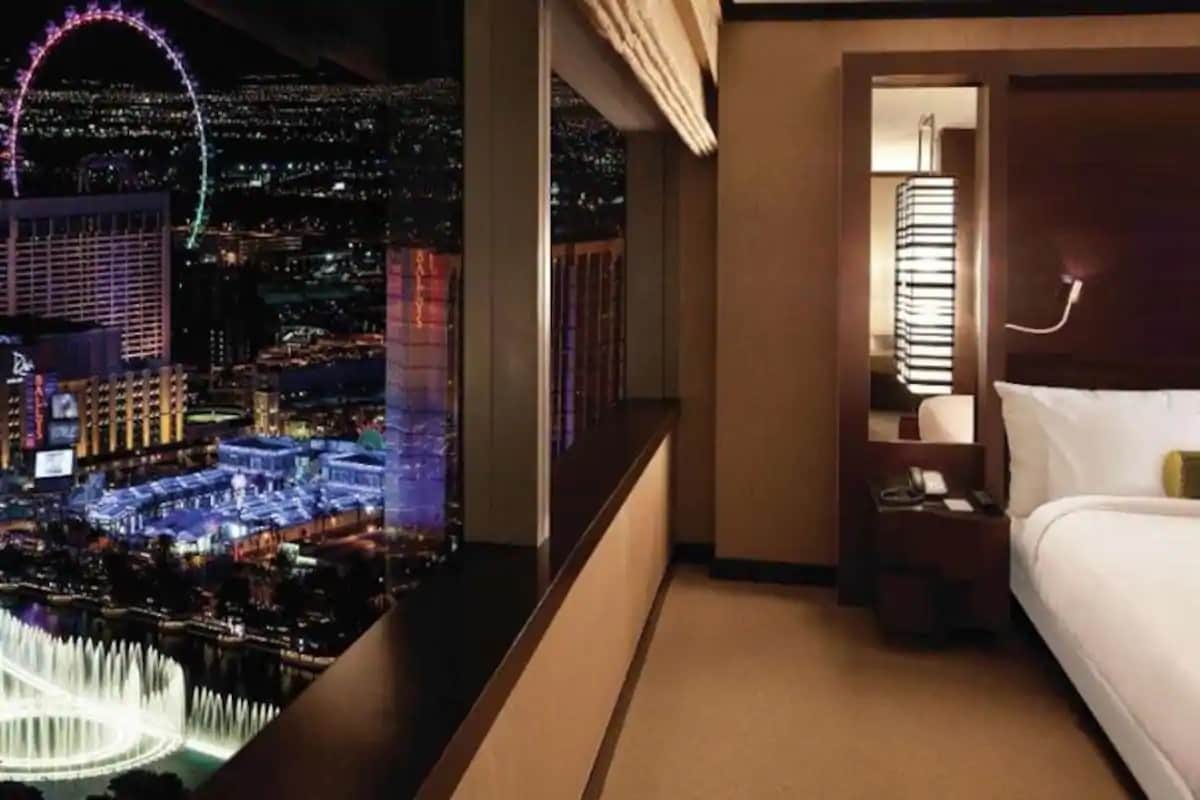 The room features a large window offering a stunning view of the Las Vegas Strip, including the colorful lights of the High Roller and the Bellagio Fountains below. A spacious king-size bed with crisp white linens is positioned against a modern accent wall.