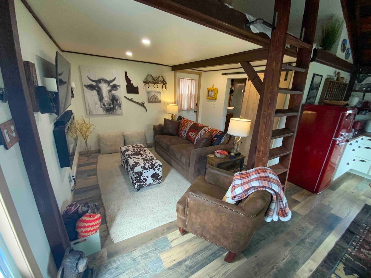 A cozy living area is presented with a spacious sofa, accent chairs, and a soft area rug. Artwork featuring local themes decorates the walls. Warm lighting creates a comfortable ambiance, while a vintage red refrigerator adds a pop of color to the space.