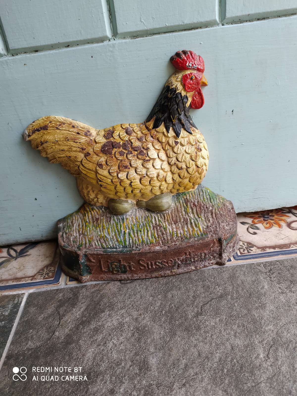 A decorative chicken figure, painted in shades of yellow and red, stands against a blue wall. The figure rests on a base labeled 'Light Sussex Hen,' complementing the surrounding patterned tiled floor.
