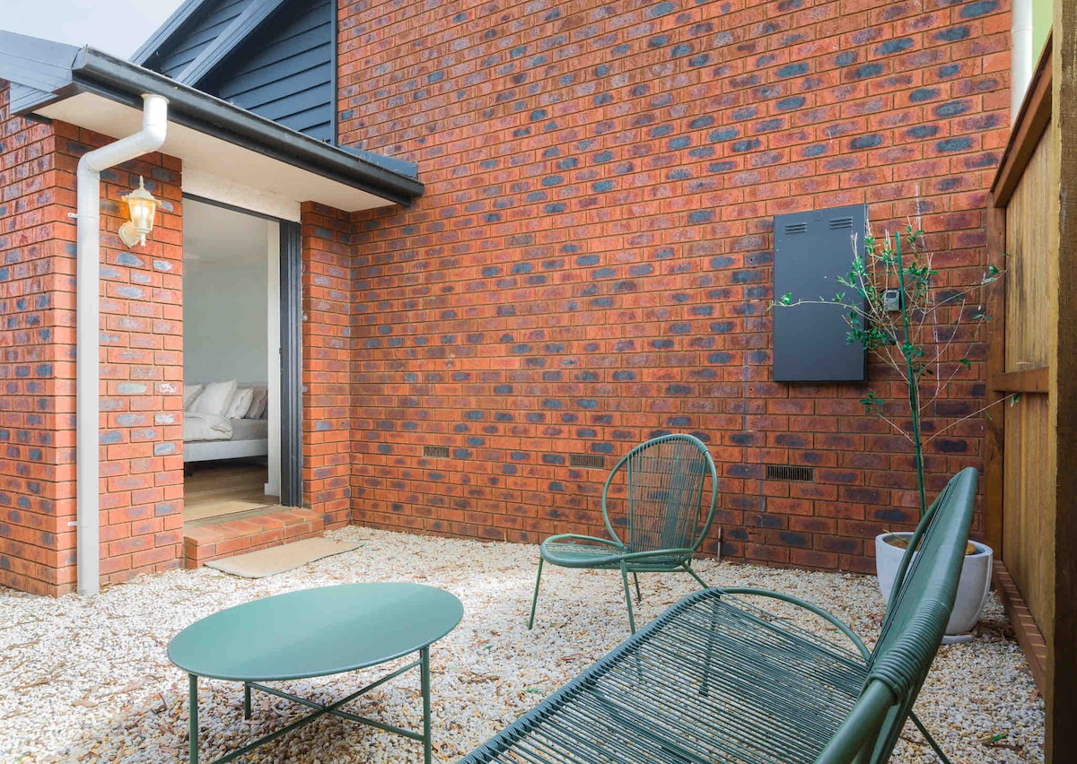 A private patio area is shown, featuring a green metal table and two matching lounge chairs. The surrounding pebbled ground complements the textured red brick wall of the guest suite. An entrance leads into the interior space, partially visible in the background.