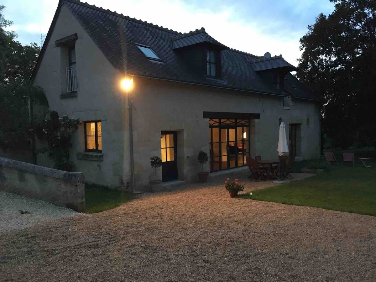 The charming exterior of a restored farmhouse is illuminated by outdoor lights during twilight. The gravel pathway leads to an inviting entrance, framed by large windows. A patio area with a dining table and chairs offers a view of the surrounding garden.