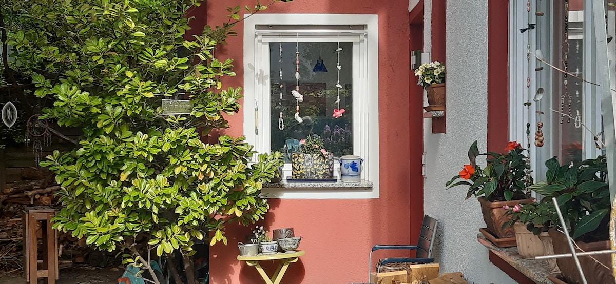 A cozy outdoor area features a window adorned with hanging decorations and flower pots. Green foliage frames the space, while a small table and a chair provide seating. The vibrant colors of the surrounding plants create a welcoming environment.