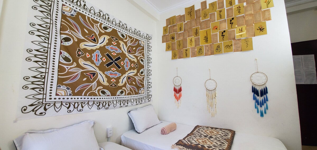 A spacious room features two beds, each adorned with crisp linens. An intricately designed wall tapestry adds vibrant patterns. Above, decorative tiles showcase a range of earthy colors and designs. Wall hangings with feathers and beads enhance the peaceful ambiance.