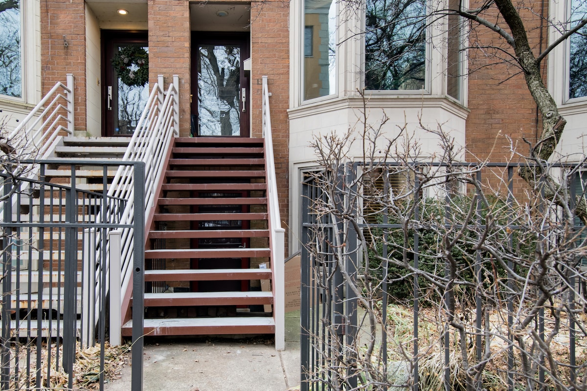 The entrance features a set of wooden stairs leading to double doors, flanked by large windows. A wrought iron railing provides support on either side, while shrubbery borders the walkway, adding a touch of greenery to the exterior.