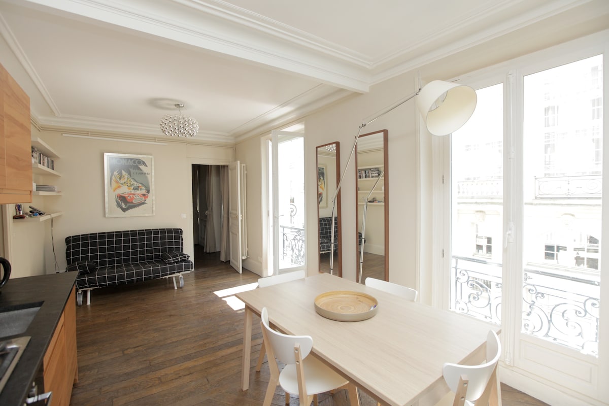 The well-lit living area features a dining table with a wooden surface and white chairs. A black-and-white checked sofa occupies one corner, while large floor-to-ceiling windows provide natural light and views of the balcony. Sleek cabinetry is attached to the kitchen area in the background.