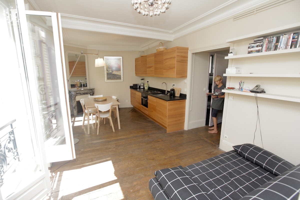 The living area features an open layout with sleek wooden cabinetry and modern appliances in the kitchen. A dining table with wooden chairs complements the space. Natural light floods through the floor-to-ceiling windows, highlighting the hardwood floors and contemporary furnishings.