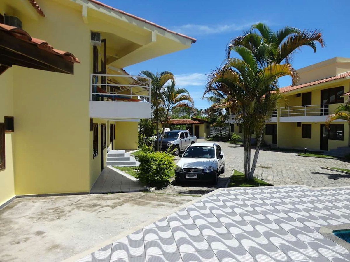 The image captures a serene outdoor view of the condominium, featuring yellow buildings surrounded by palm trees. A car is parked nearby, and the patterned pavement leads towards the pool area, providing a glimpse of the inviting landscape.