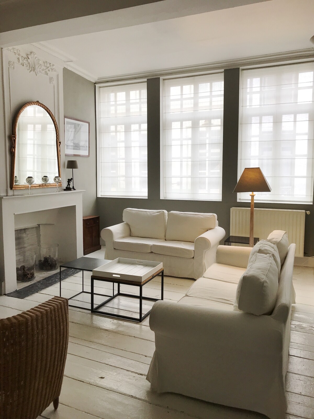 A bright living area features white sofas arranged around a glass coffee table, with large windows allowing natural light to fill the space. A mirror and a lamp create a cozy ambiance, while a decorative fireplace adds subtle character to the room.