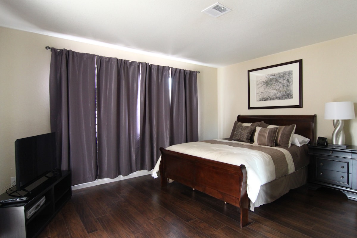 A tastefully furnished bedroom features a spacious bed with neutral-toned bedding. Dark curtains frame the window, allowing filtered light. A sleek nightstand with a lamp complements a flat-screen tv positioned across from the bed, enhancing the room's functionality.