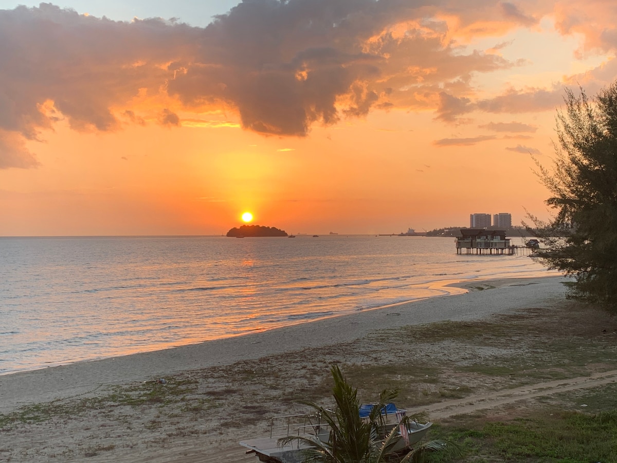 A serene sunset is captured over a calm sea, with the sun descending on the horizon. A distant island is visible, alongside silhouettes of boats and buildings along the shoreline. The soft colors of the sky blend gently with the water's surface, creating a tranquil scene.