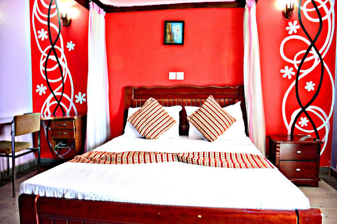 A vibrant bedroom features a wooden bed with decorative pillows, surrounded by painted walls in bold red and white patterns. Two bedside tables are positioned next to the bed, with soft lighting provided by wall sconces. A small artwork hangs above the bed, adding an artistic touch.