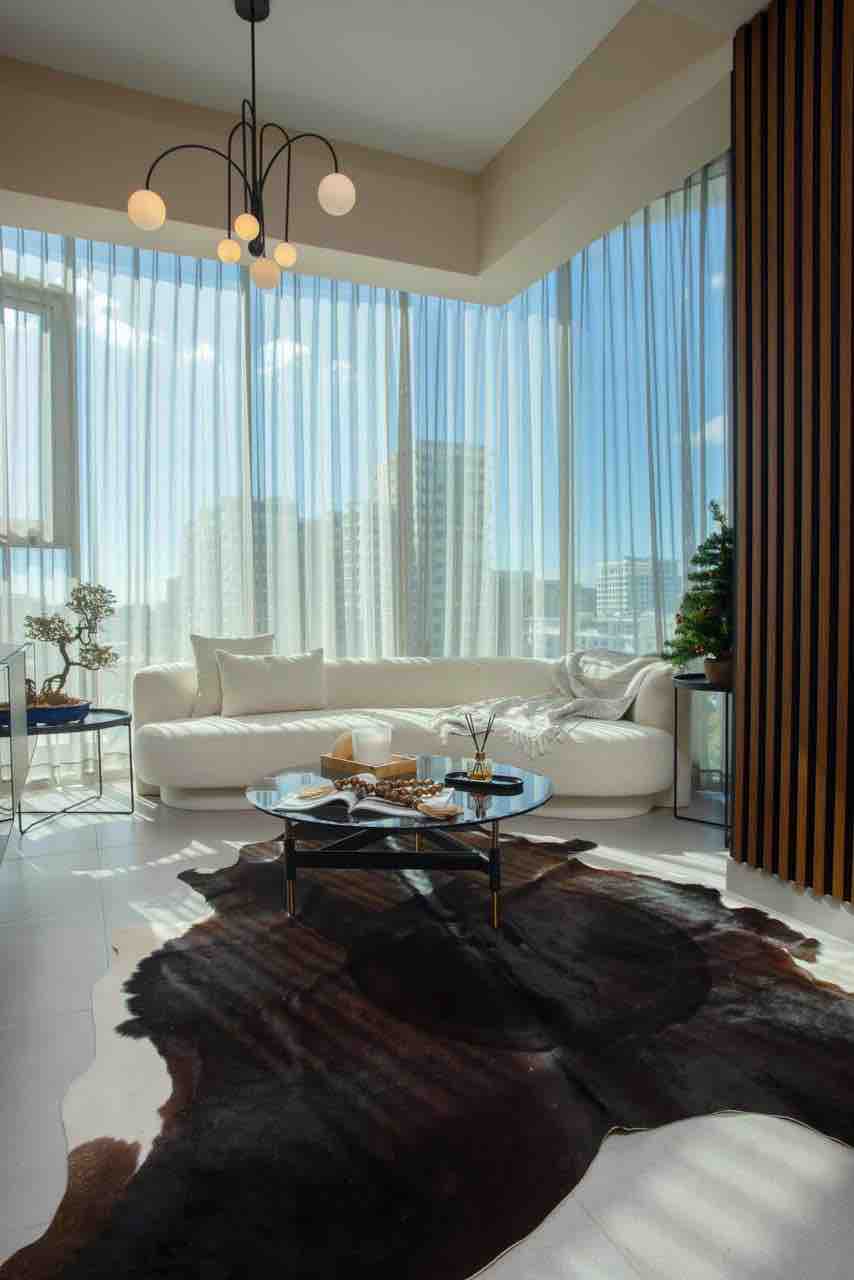 A sunlit living area features large floor-to-ceiling windows that offer city views. A round black coffee table is centered on a textured rug, complemented by a light-colored sofa adorned with cushions. Decorative plants are placed strategically, enhancing the space's minimalist design.