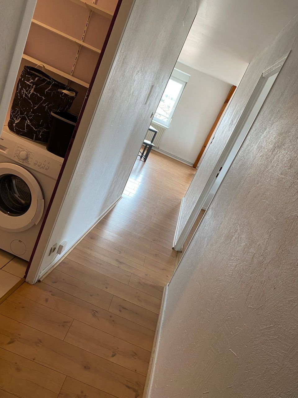 A hallway is depicted, featuring light wood flooring that enhances the spaciousness. A washing machine is visible on the left, with a doorway leading to a room at the far end. Natural light streams in through a window, illuminating the neutral-colored walls.