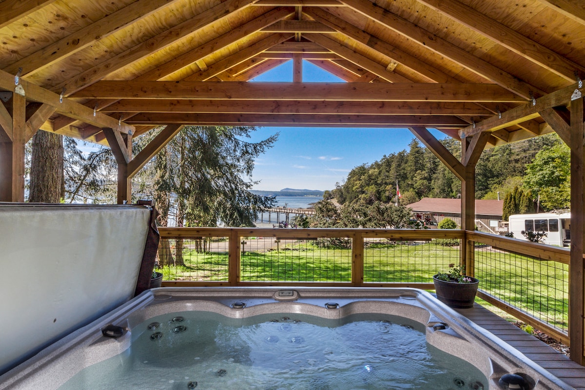 A covered hot tub area features wooden beams and a clear view of the water and landscape. The hot tub is filled and surrounded by a wood deck, with a planter nearby, creating a peaceful atmosphere for relaxation and enjoyment of the scenery.