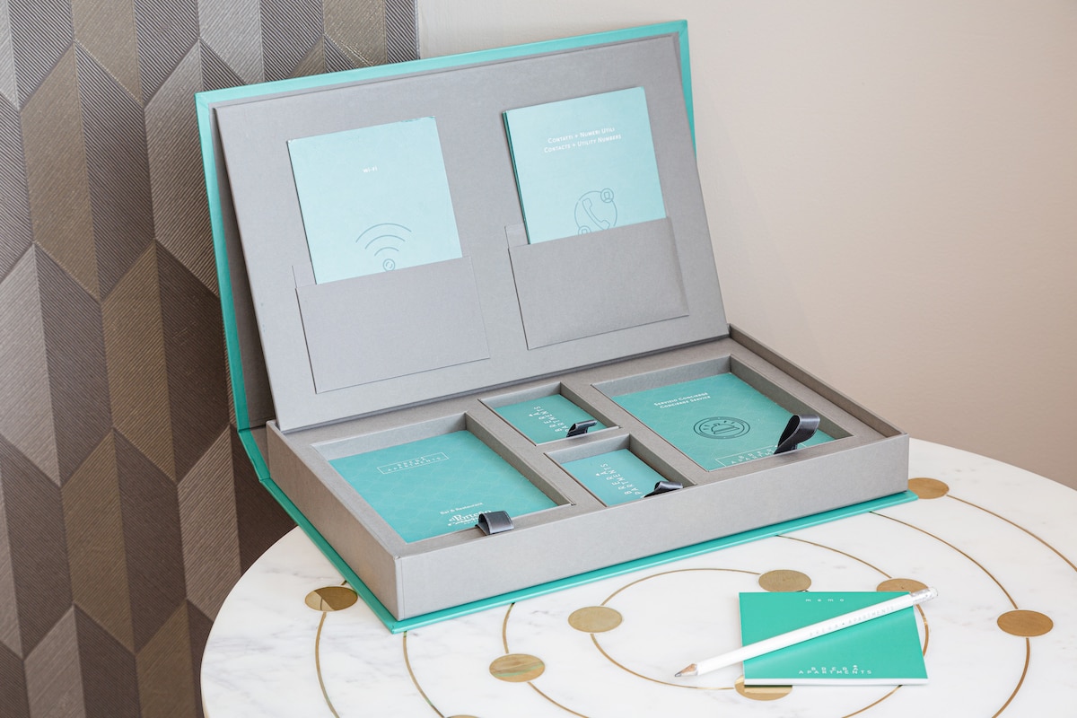A neatly arranged welcome kit is displayed on a round marble table. The kit features a grey box with turquoise accents, containing booklets and cards that provide information on amenities. A pen rests beside the kit, complementing the organized appearance.