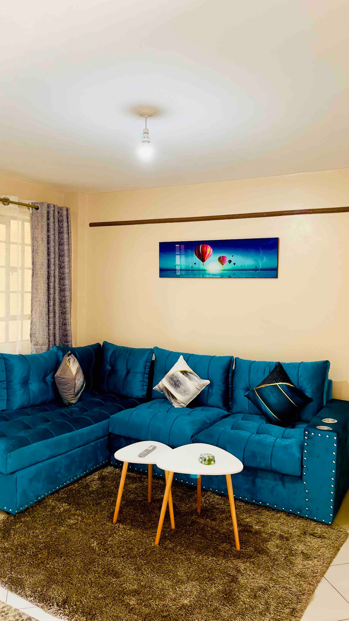 A modern living area features a large, turquoise sectional sofa adorned with decorative pillows. A minimalist white coffee table sits between two wooden legs on a plush area rug. A piece of artwork with colorful hot air balloons hangs on the wall, complemented by natural light from a window.
