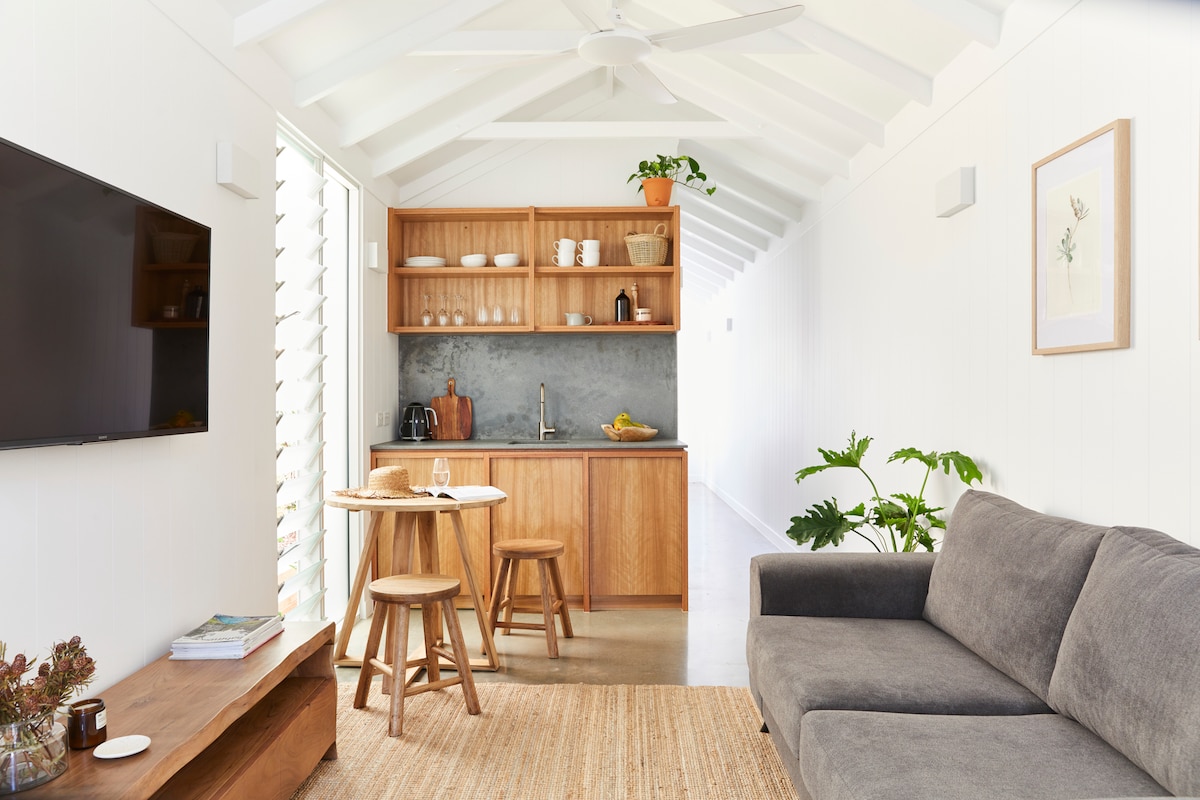 An open living area showcases light-colored walls and a high ceiling with exposed beams. A comfortable gray sofa is placed beside a small wooden table and two stools. The kitchenette features wooden cabinetry and essential appliances, with natural light pouring in from large windows.