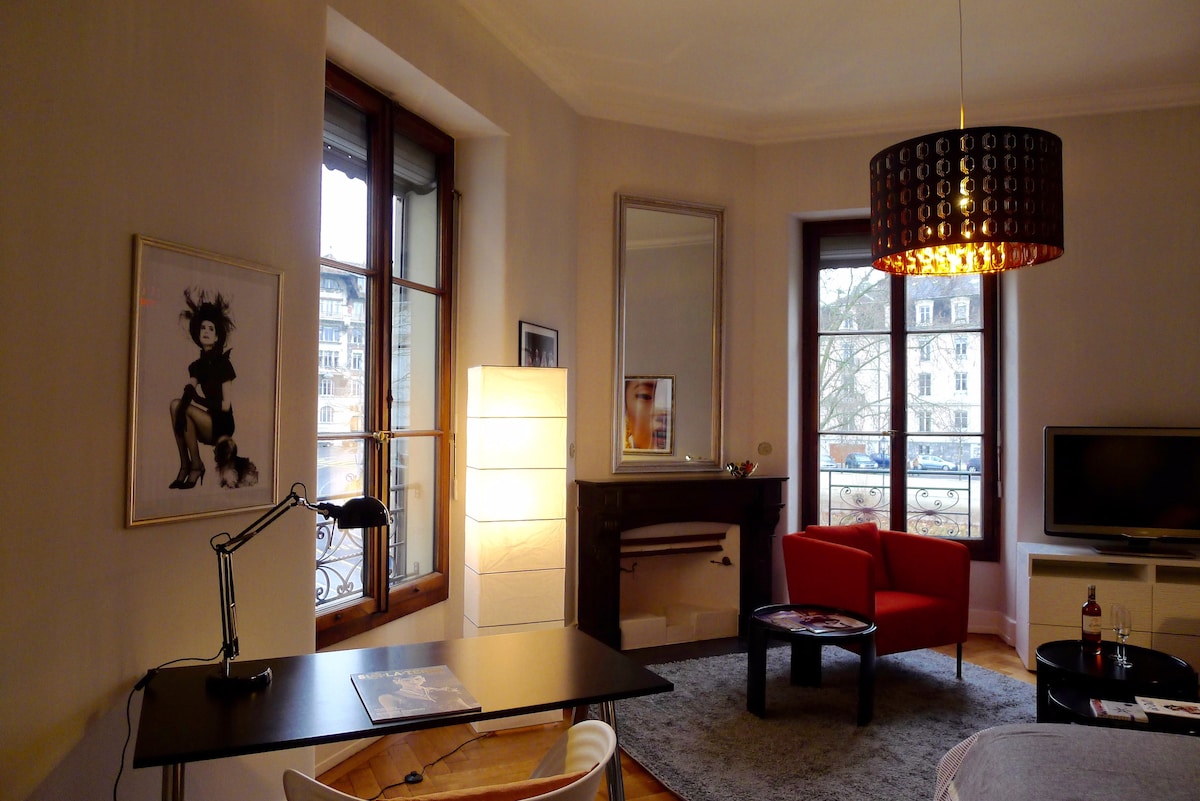 A stylish living area is presented, featuring a contemporary desk with a desk lamp and a comfortable red armchair. A light fixture hangs above, complementing the large windows with views of the street outside. A mirror and TV also enhance the room's appeal.