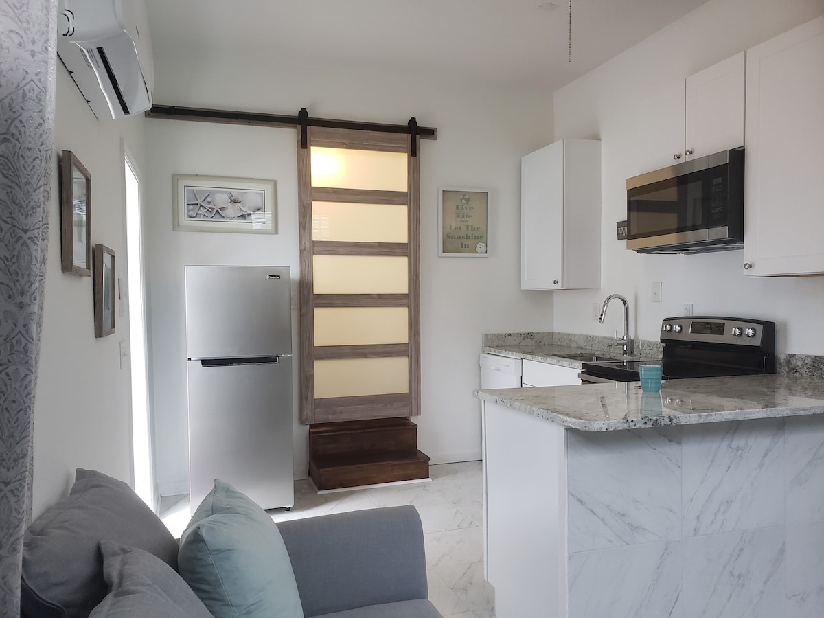 A modern kitchen area includes a sleek refrigerator and microwave, with marble countertops providing a clean and functional space. A barn-style sliding door adds character, leading toward a stylish entryway. Natural light fills the room, complemented by soft decorative touches.