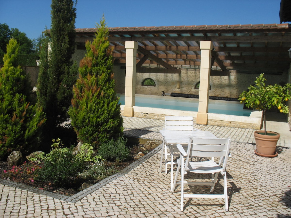A serene outdoor space features a stone-paved area with white lounge chairs placed nearby. Lush green hedges and flowering plants create a natural border around the area, which overlooks a swimming pool that is partially sheltered by a wooden roof structure.