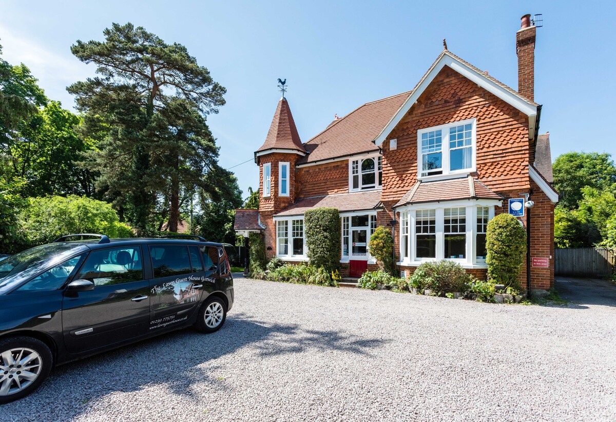 A charming Victorian-style guest house is set amidst lush greenery. The brick exterior features large windows and a decorative turret. A gravel driveway accommodates a parked vehicle, while manicured hedges add to the inviting entrance.