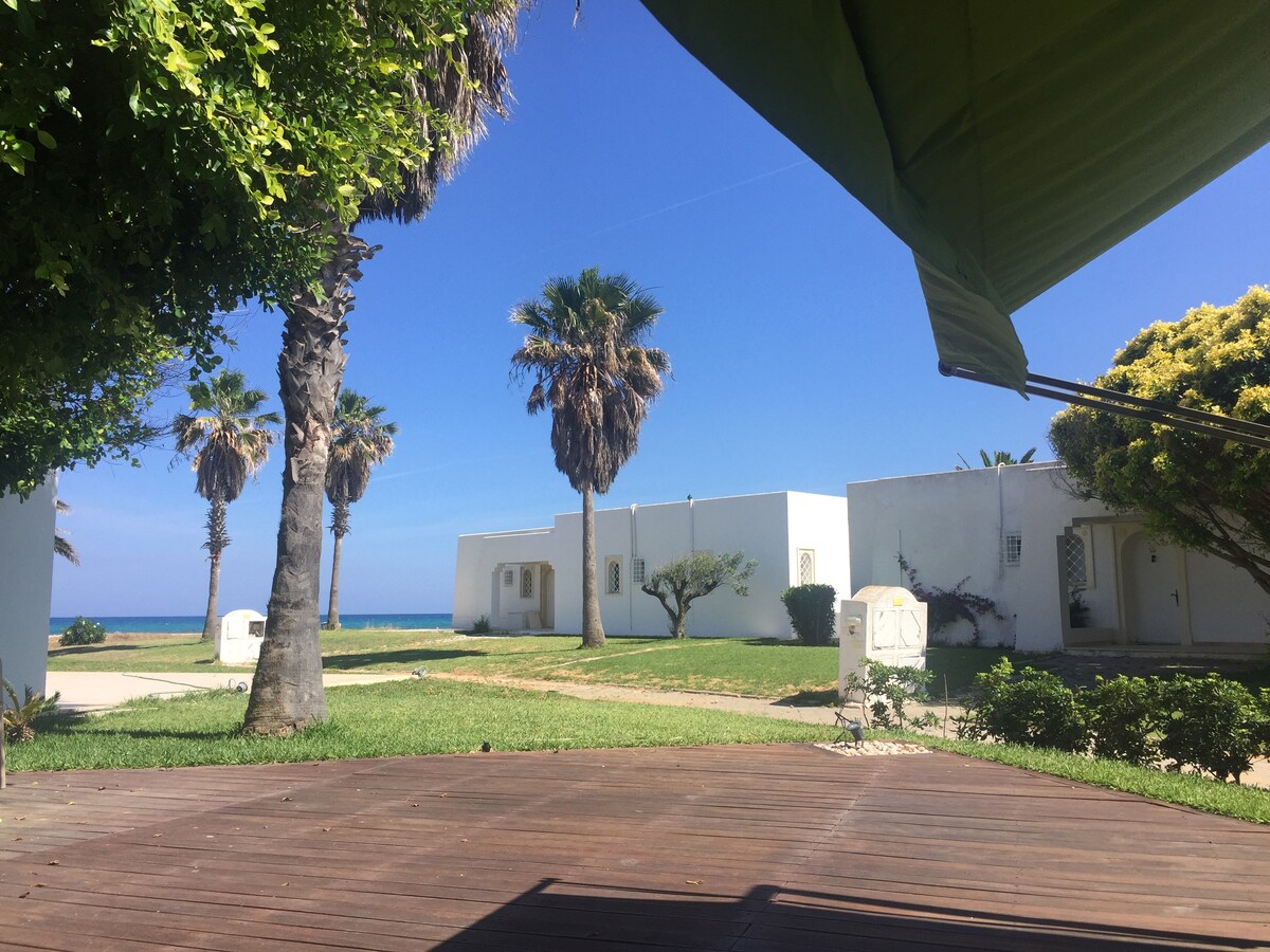 A view of a well-maintained lawn featuring palm trees and lush greenery. White bungalows are visible in the background against a clear blue sky, with the sea seen in the distance. The outdoor area is marked by a wooden deck, providing a tranquil space for relaxation.