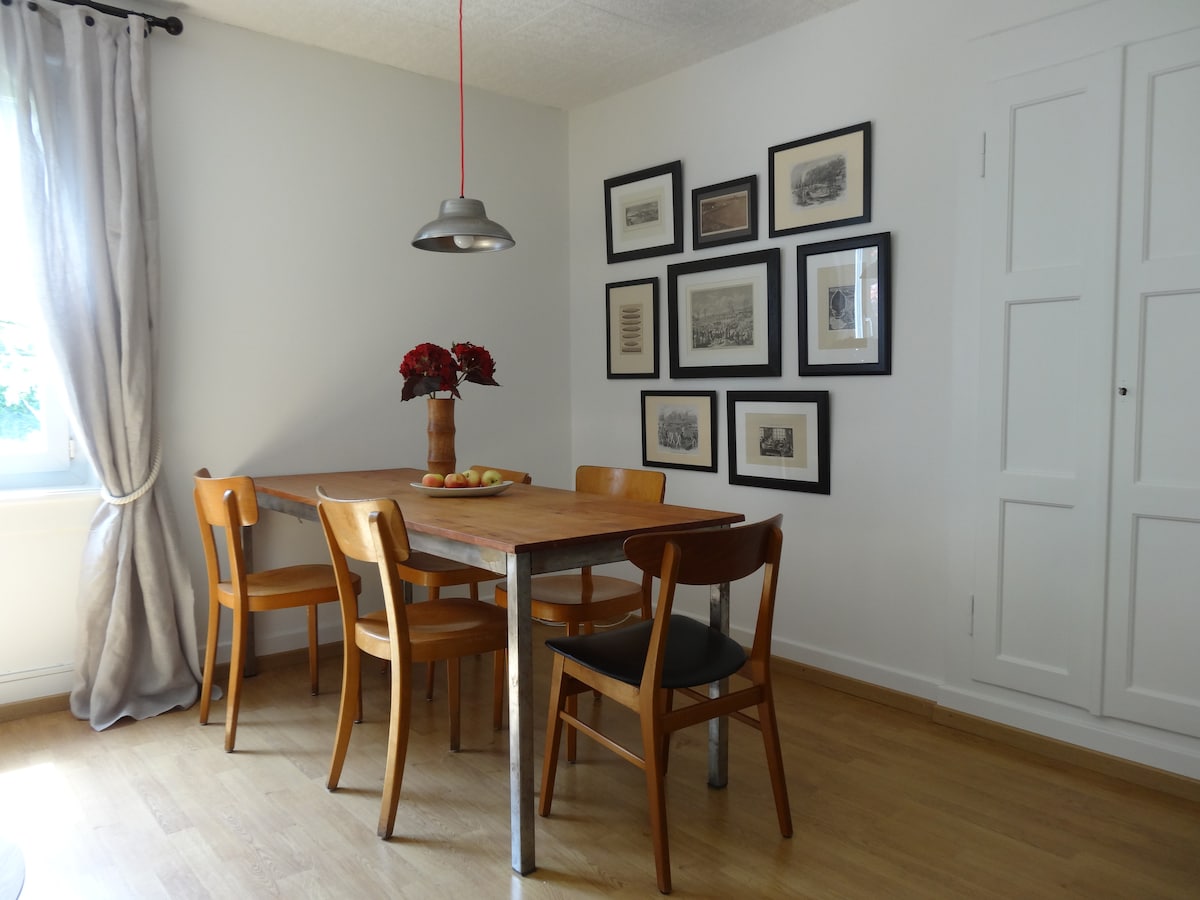 A dining area is displayed with a wooden table that accommodates six, surrounded by six chairs. Above, a pendant light offers soft illumination. A bouquet of flowers is center on the table, while framed art is arranged on the wall, adding subtle character to the space.