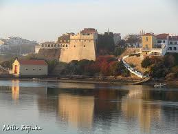 A historic fortress is situated atop a hill, overlooking a calm body of water that reflects the structure and nearby buildings. Surrounding greenery and autumn colors are visible, creating a tranquil view of the village and landscape.