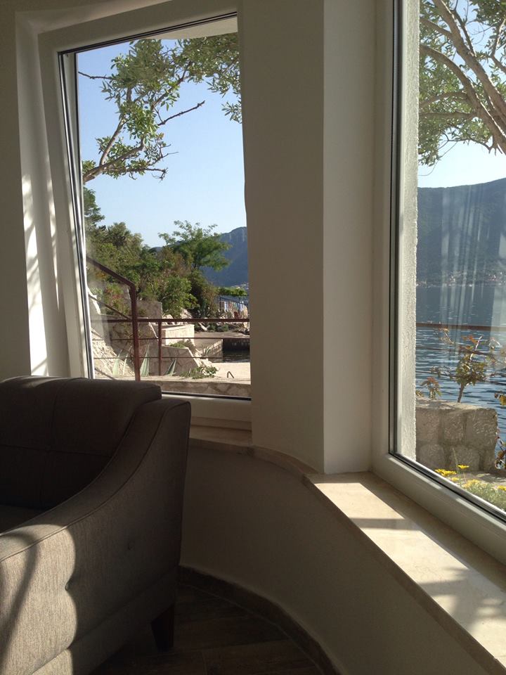 A large window with clear glass offers a view of the serene sea and surrounding greenery. Sunlight casts gentle shadows, and a comfortable gray armchair is positioned nearby, creating a relaxing space to enjoy the natural scenery.