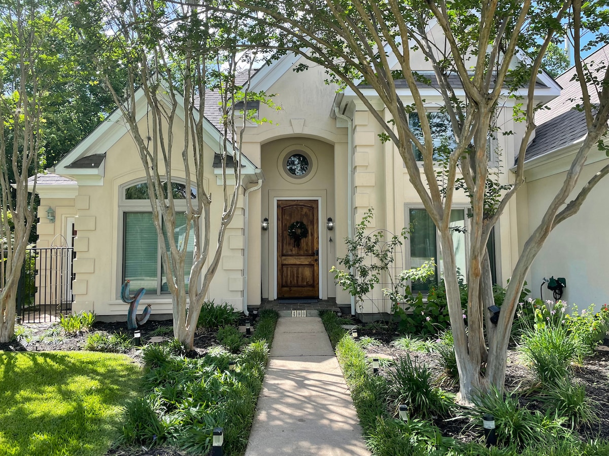 The entrance of a custom-built home presents an elegant oversized oak front door centered beneath a decorative window. Flanking the path are well-kept gardens featuring various shrubs and small trees, creating a welcoming ambiance.