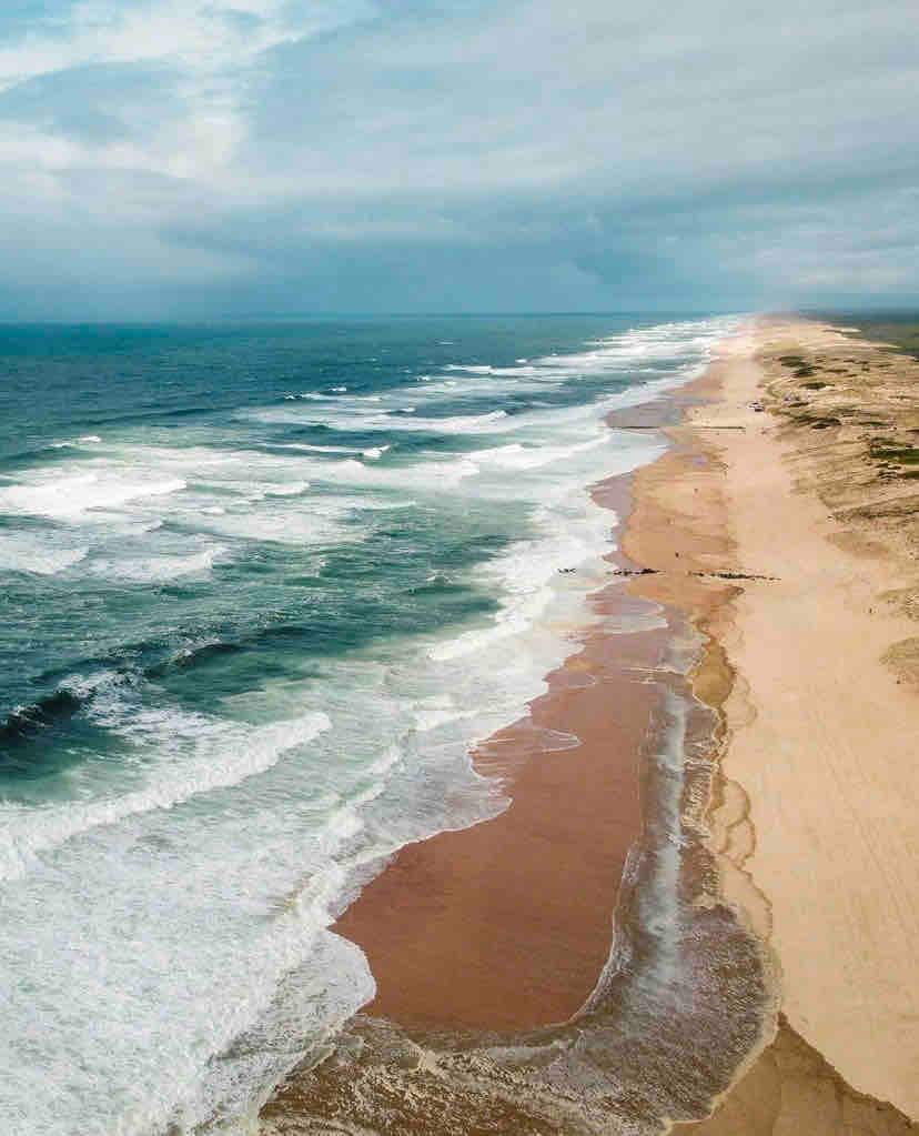 A wide view of a beach landscape reveals soft, sandy stretches meeting rolling waves. The sea showcases various shades of blue and green, with white frothy crests. Cloudy skies above create a calm atmosphere, while the shoreline extends into the distance.