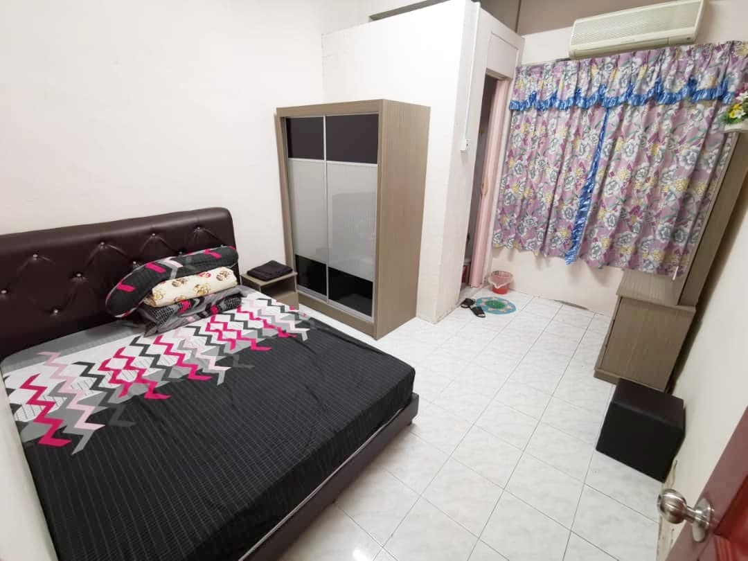 The sleeping area features a spacious bed with a patterned bedspread. A wardrobe stands against one wall, while a small side table is positioned nearby. Colorful curtains frame a window that allows natural light into the room, which has a clean tiled floor.