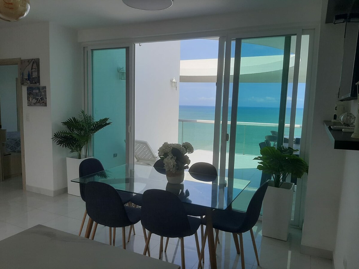 A dining area features a glass table surrounded by six dark chairs. Natural light fills the space through large sliding glass doors that lead to a balcony overlooking the sea. Green plants are placed in white pots, adding a touch of nature.