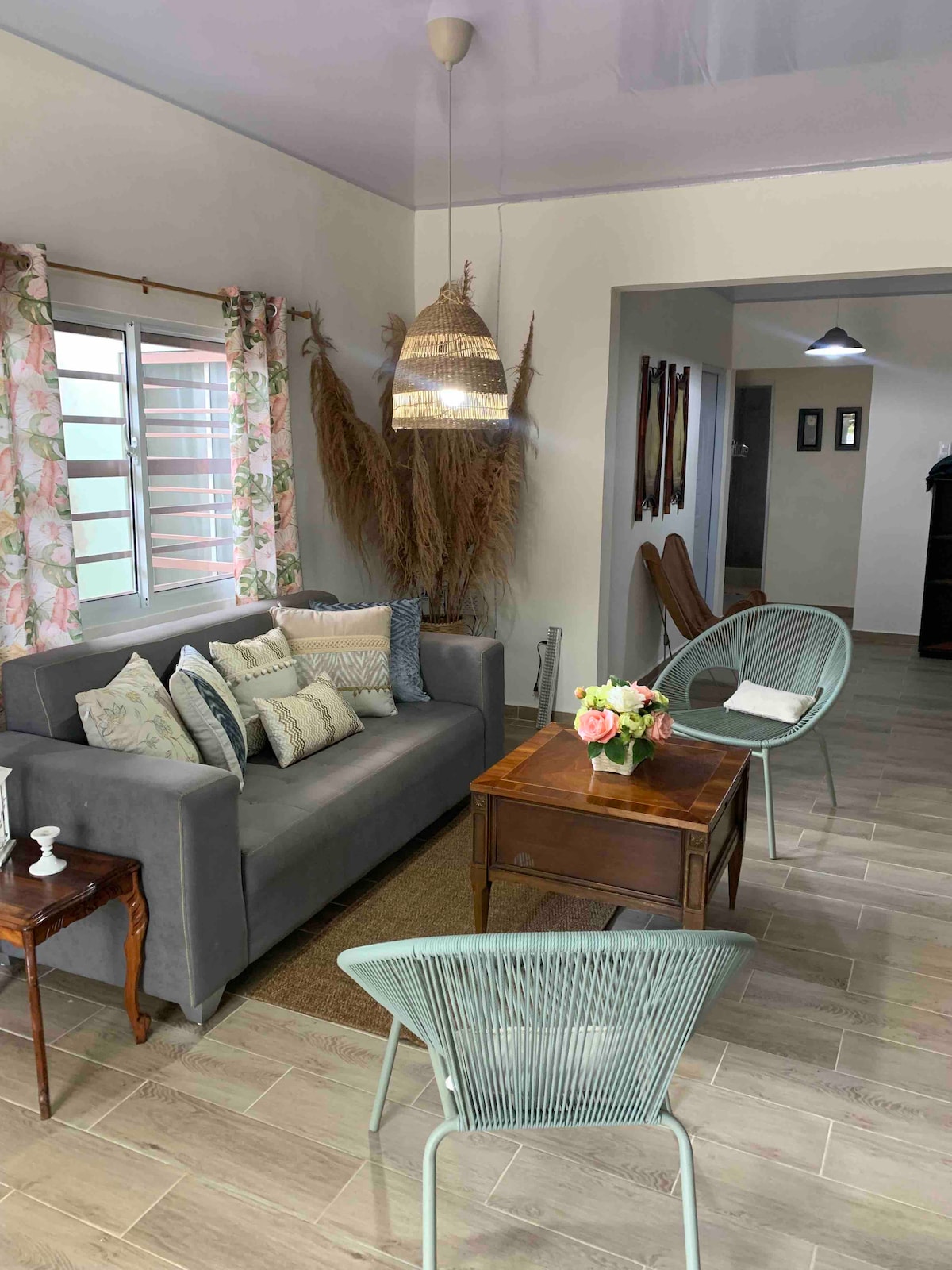 A spacious living area is furnished with a comfortable gray sofa adorned with decorative pillows. A wooden coffee table sits in the center, accompanied by two light green chairs. Natural light enters through a window with floral curtains, enhancing the serene ambiance.
