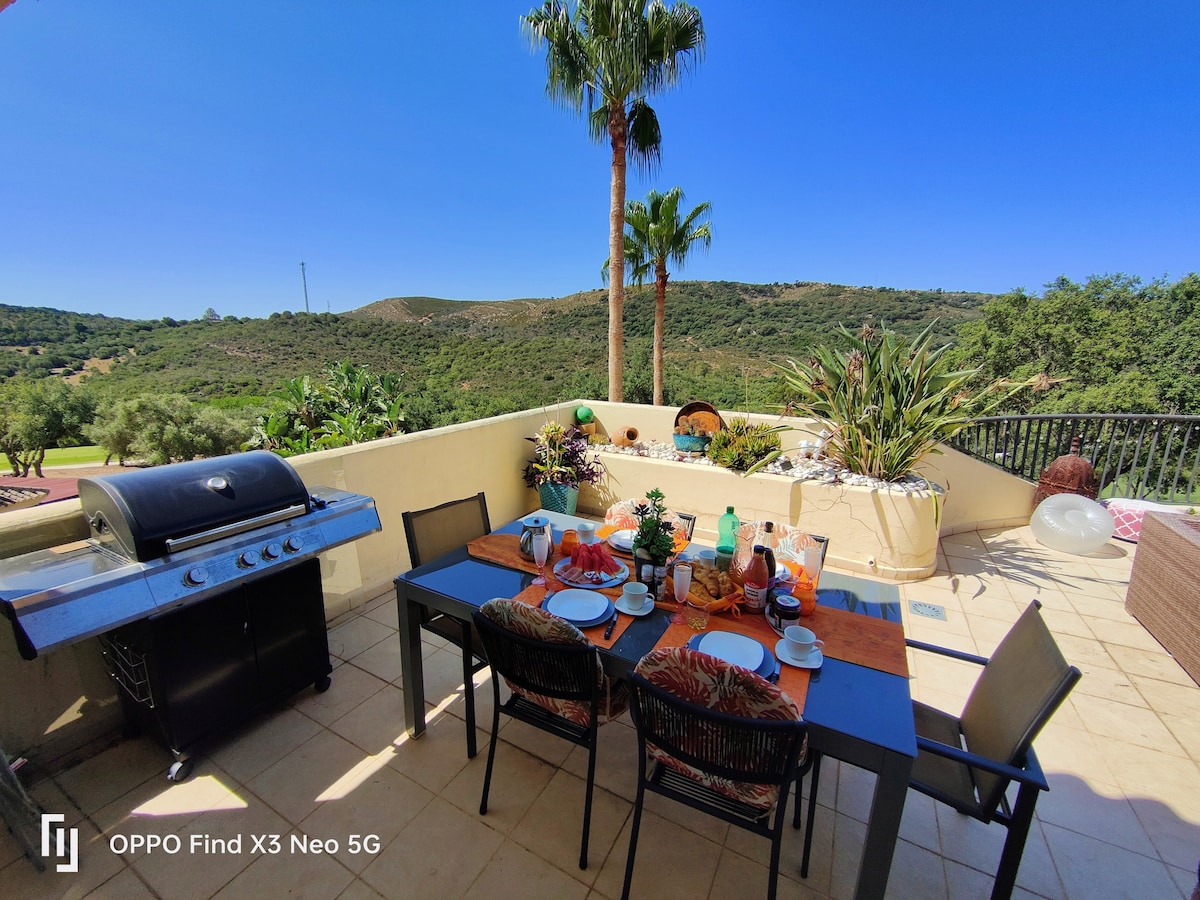 A spacious terrace features a dining table set with plates and glasses, paired with comfortable chairs. A barbecue grill is positioned nearby, surrounded by diverse greenery and mountain views under a clear blue sky. The bright outdoor space invites relaxation and al fresco dining.